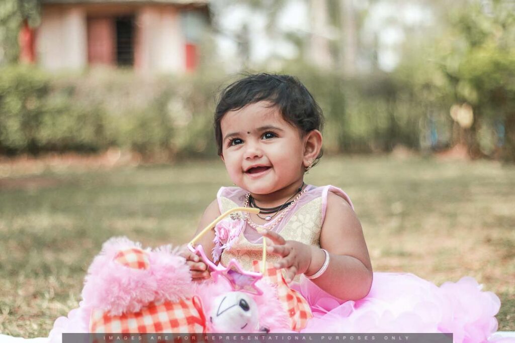 Smiling baby girl in a pink dress sitting outdoors with soft toys.
