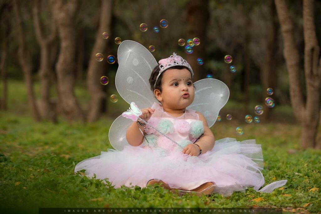 Baby dressed as a fairy with wings and a tiara, sitting on green grass surrounded by bubbles in a forest-like setting.